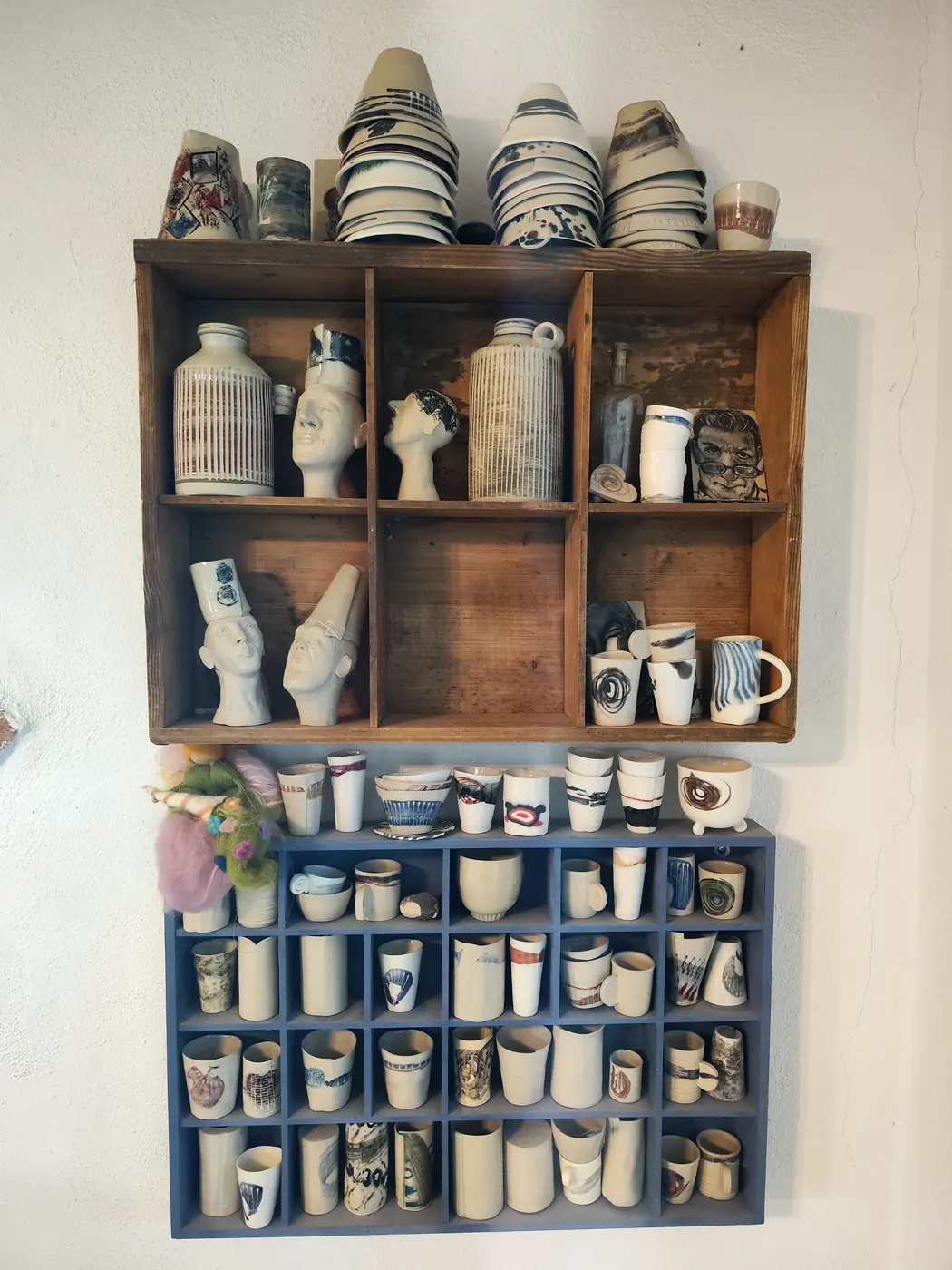 Wandregal aus altem Holz und ein blauer Setzkasten voller Becher, oben gestapelte Schalen, im Regal modellierte Köpfe